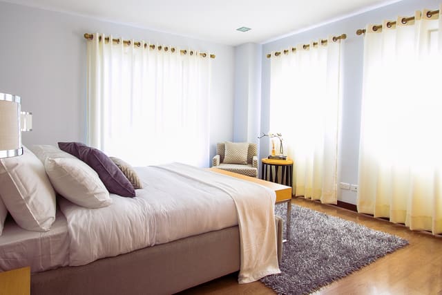Bedroom with a made bed, purple accent pillows, a gray rug on wood floors and tall windows with sheer curtains.