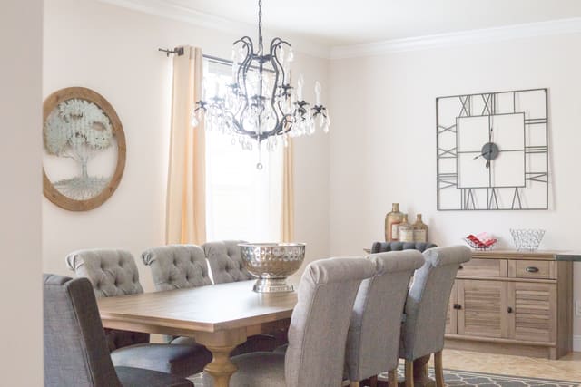 Dining room with a wood table, gray upholstered chairs, a crystal chandelier and a window with sheer curtains.