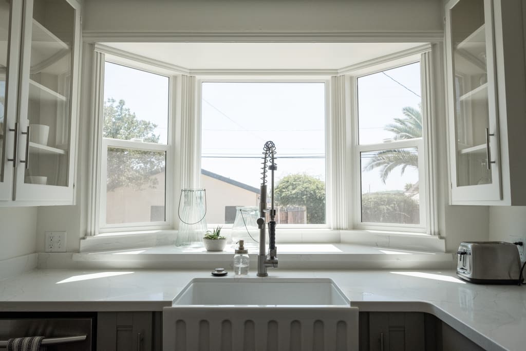 Bright kitchen with a farmhouse sink, stone counters and glass-front cabinets beside a bay window.